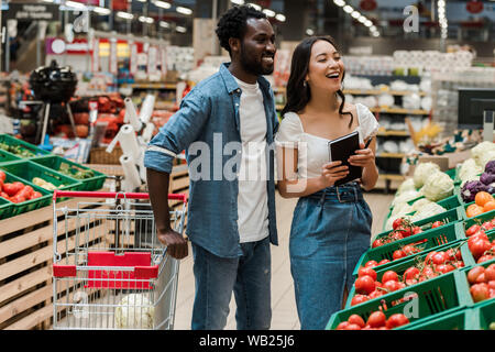 Fröhlich afrikanische amerikanische Mann in der Nähe von attraktiven asiatischen Frau mit Notebook im Supermarkt Stockfoto