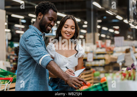 Selektiver Fokus von Happy african american man berühren rote Tomaten in der Nähe von asiatischen Frau in Store Stockfoto