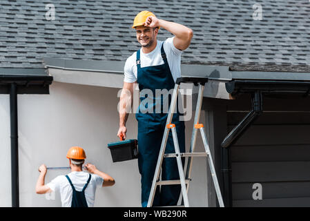 Selektiver Fokus der glücklichen Handwerker berühren, Helm und stehend mit Toolbox auf Leiter in der Nähe von Kollegen Stockfoto