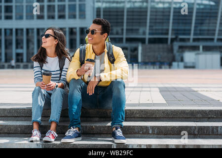 Interracial Touristen mit Rucksäcken auf Treppen mit Papier Tassen Kaffee und Wegsehen Stockfoto