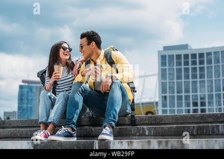 Interracial Freunde sitzen auf der Treppe und einander mit Rucksäcken und Tassen Kaffee Stockfoto