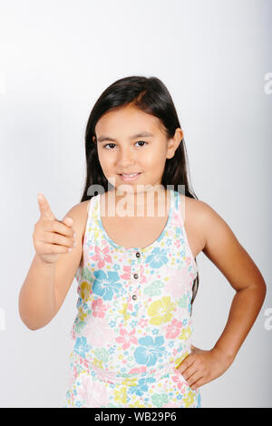 Ein 9-jähriges Mädchen ist auf Kamera. Sie steht vor einer weißen Wand. Sie lächelt. Sie hat lange dunkle Haare. Sie hat zwei Funktionen. Sh Stockfoto