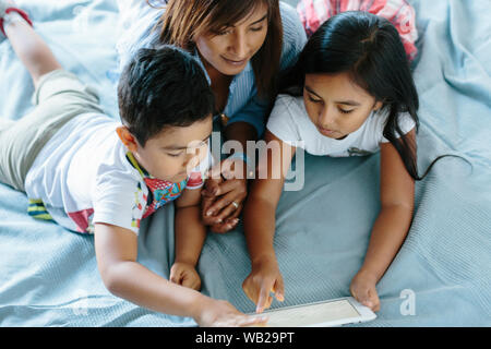 Eine Familie mit einem 9 Jahre alten Mädchen, ein 5-Jähriger Junge und ihre Mutter auf dem Bett liegend sind. Sie sind auf der Suche nach einem Grafiktablett. Sie sind mit dem Sommer kleiden gekleidet Stockfoto