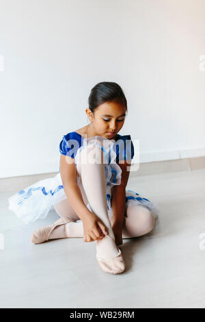 Ein 9-jähriges Mädchen sitzen auf dem Boden. Sie setzen ihr Ballett Schuhe an. Sie trägt ihr Ballerina Outfit. Sie hat Hispanic Ethnizität Features. Stockfoto