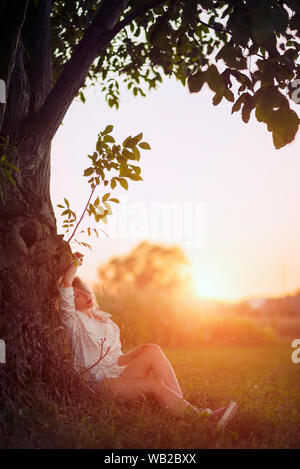Junge Frau sitzt unter einem Baum bei Sonnenuntergang Stockfoto
