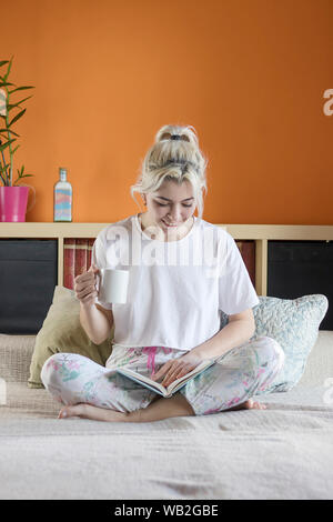 Blonde hübsche Dame im Schlafanzug ist trinken Ihren Kaffee am Morgen sitzen auf dem Bett Stockfoto