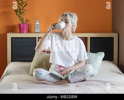 Blonde hübsche Dame im Schlafanzug ist trinken Ihren Kaffee am Morgen sitzen auf dem Bett Stockfoto