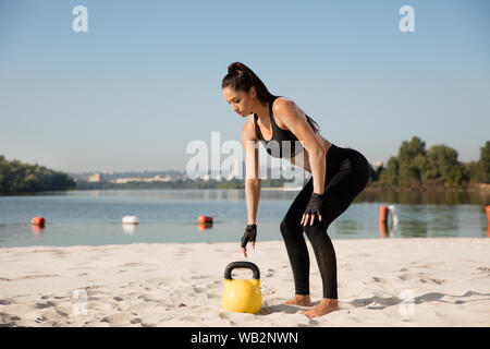 Junge gesunde Frau hockt mit Gewichten an den Strand. Einzelnen kaukasischen weibliche Modell üben an der Flussseite in sonniger Tag. Konzept der gesunden Lebensstil, Sport, Fitness, Bodybuilding. Stockfoto