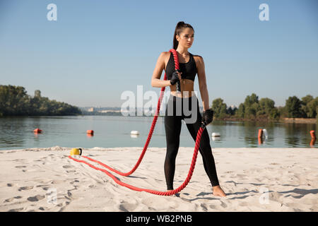 Gesunde junge Frau mit der Seile an den Strand posieren. Einzelnen kaukasischen weibliche Modell üben an der Flussseite in sonniger Tag. Konzept der gesunden Lebensstil, Sport, Fitness, Bodybuilding. Stockfoto