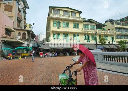 Eine alte Frau mit einem Kinderwagen geht eine große restaurierte Gebäude bei Saphan Han, ein KLONG (Kanal) Brücke in Chinatown/Pahurat, Bangkok, Thailand Stockfoto