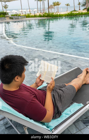 Asiatischer mann lesen Buch beim Entspannen am Pool Stockfoto