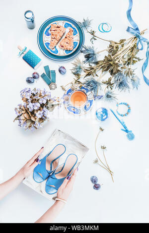 Frau Hände halten neue blaue Schuhe in Feld auf weißen Desktop. Feminine Stillleben mit kosmetischen Produkten, Blumen Bündel, Tasse Kaffee mit Kuchen Stücke, j Stockfoto