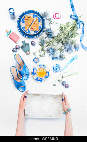 Frau Hände halten leere Box auf weißen Desktop mit neuen blauen Sandalen und feminin noch Leben von kosmetischen Produkten, Blumen Bündel, Tasse Kaffee mit Kuchen Stockfoto