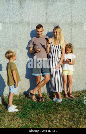 Foto von Frau, Mann und Tochter mit Telefone in ihren Händen und Sohn neben Betonwand auf der Straße im Sommer Tag Stockfoto