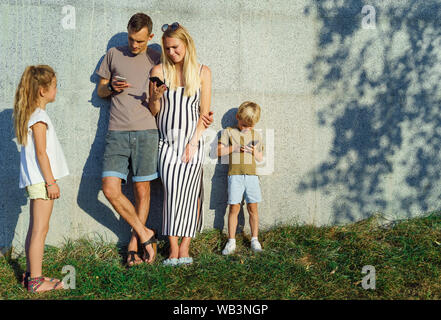 Bild von Frau, Mann und Sohn mit Telefonen in den Händen und Töchter neben Beton Wand draußen auf Sommer Tag Stockfoto