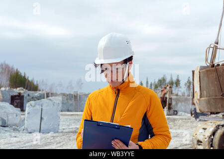 Weibliche Geologe oder ein bergbauingenieur Schreibt etwas in einer Karte - Fall mitten in einem Steinbruch mit Baumaschinen Stockfoto
