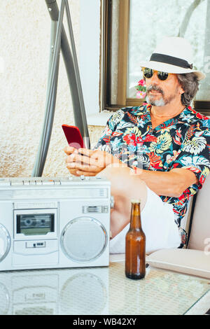 Reifer Mann auf sein Handy zu schielen, während auf einer Schaukel sitzend Stuhl, mit einem Bier und Musik aus einem Radio-/Kassetten-Player Stockfoto