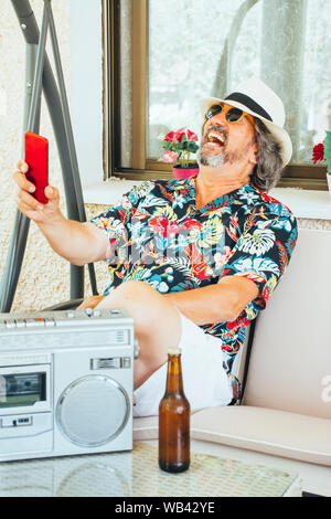Reifer Mann Spaß mit seinem Handy, während auf einer Schaukel sitzend Stuhl mit einem Bier und Musik aus einem Radio-/Kassetten-Player Stockfoto