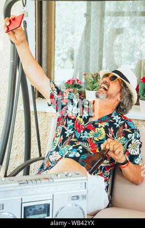 Reifer Mann genießen Unter selfies mit Handy beim Sitzen auf einem Stuhl, mit einem Bier und Musik aus einem Radio-/Kassetten-Player Stockfoto
