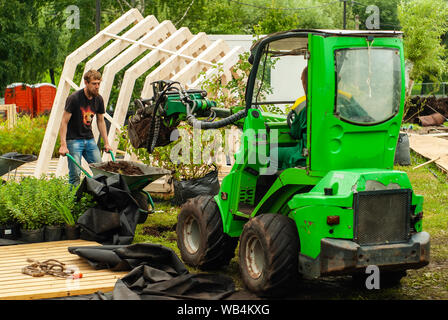 Perm, Russland - Juli 12, 2019: zwei Arbeiter den Boden graben mit Hilfe einer Schubkarre und ein Mini Bagger bei der Arbeit auf dekorative lanscape Stockfoto