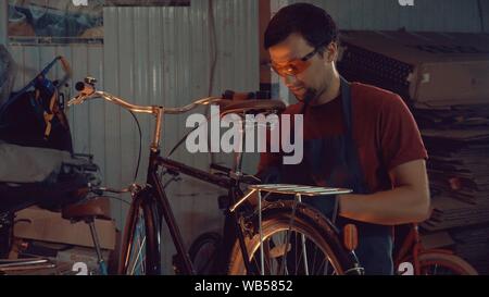 Thema Small Business Fahrrad reparieren. Eine junge Kaukasier brünett Mann tragen Sie eine Schutzbrille, Handschuhe und Schürze verwendet eine hand Tool zum Reparieren und einstellen Stockfoto