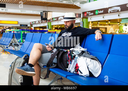 Junger Mann mit Hut mit einem Smartphone während wartet mit seinem Gepäck in einem Flughafen Stockfoto