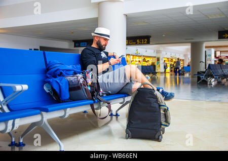 Junger Mann mit Hut mit einem Smartphone während wartet mit seinem Gepäck in einem Flughafen Stockfoto