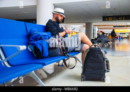 Junger Mann mit Hut mit einem Smartphone während wartet mit seinem Gepäck in einem Flughafen Stockfoto