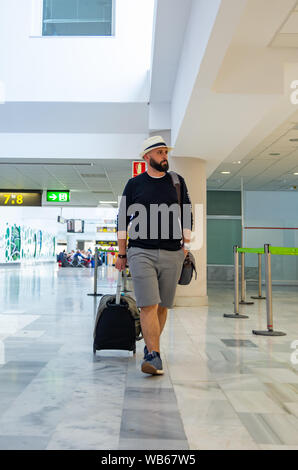 Junger Mann mit Hut gehen durch einen Flughafen Terminal mit einem Koffer Stockfoto