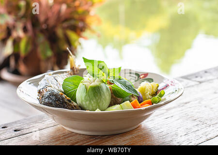 Gebratener Reis mit Chili würzig mit Fisch und Gemüse in Weiß Platte auf dem Holztisch. Hintergrund Flüsse und Bäume. Stockfoto
