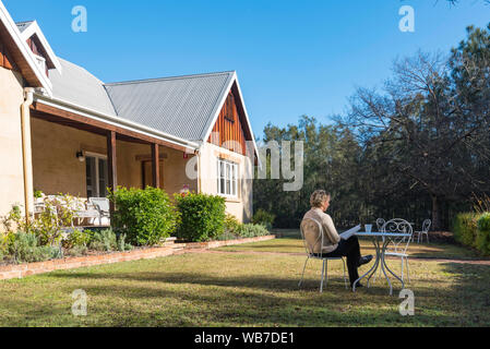 Eine mittlere gealterte weiße Frau sitzt an einer weißen outdoor Einstellung in morgen Winter Sonnenlicht in einem Garten vor einem Haus mit einer Tasse Tee ein Buch lesen Stockfoto
