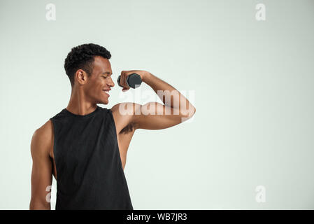 Die perfekten Körper. Muskulösen jungen afrikanischen Mann Training mit Hanteln und lächelnd, während gegen grauen Hintergrund stehen. Studio gedreht. Sport Konzept Stockfoto