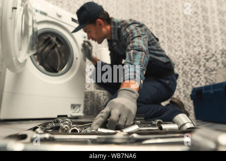 Porträt der jungen Handwerker in Uniform neben gebrochenen Waschmaschine und versuchen, es zu beheben. Horizontale erschossen. Von der Seite. Selektiver Fokus auf eine Hand und Toolbox Stockfoto