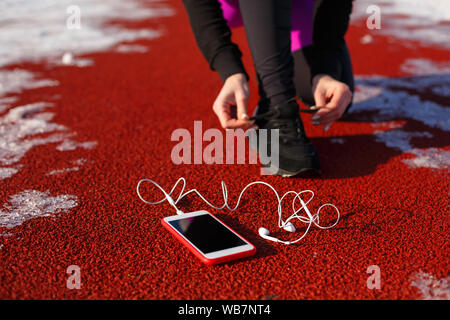 Mädchen Athlet in schwarzen Sneakers, geduckt auf der roten Spur ausgeführt werden. In der Nähe ist ein Telefon mit drahtgebundenen Kopfhörern. Kalten verschneiten Wetter. Stockfoto