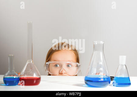 Ein kleines Mädchen in Schutzbrille versteckt sich in einem Chemielabor. Das Kind sieht aus der Tabelle. Stockfoto