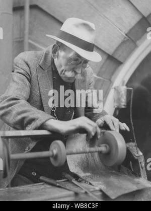 Schärfen der Messer in den 1930er Jahren. Eine reisende Messerschleifer mit einem tragbaren Schleifstein ist Schärfen eines Messers. Er hat seine Brille auf der Nase und konzentriert sich auf die Arbeit. Tropft Wasser auf den Stein aus einem kleinen oben kann Es haning. Stockfoto