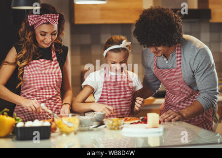 Glückliche Eltern und ihre Tochter bereiten Mahlzeit zusammen in der Küche, während kleine Mädchen, Ketchup auf der Pizza Teig. Stockfoto