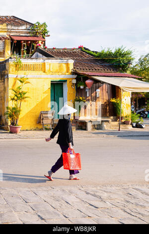 Frau mit traditionellen vietnamesischen Hut und Tasche in der Straße in der Altstadt von Hoi An in Südostasien in Vietnam am Morgen. Stadtbild mit Lady und Stockfoto