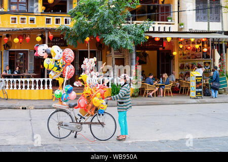 Hanoi, Vietnam - 16. Februar 2016: Stadtbild mit Frau verkaufen Spielzeug Ballons vom Fahrrad auf der Straße in der Altstadt von Hoi An, Asien, Vietnam. Menschen essen in Restaurants in Hoian auf Hintergrund Stockfoto