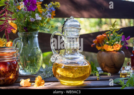 Flaschen Kräuter und ätherische Öle, die mit frischen Pflanzen und Blumen im Hintergrund Stockfoto