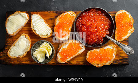 Nahaufnahme von rotem Kaviar in der Schüssel und Sandwiches auf Holzschneidebrett auf schwarzem Hintergrund. Draufsicht. Flach liegend Stockfoto