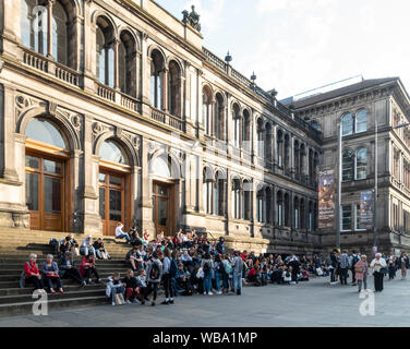 Eine grosse Gruppe von Menschen vor dem Haupteingang des National Museum of Scotland in Kammern St, Edinburgh. Erwachsene & Jugendliche sitzen auf Schritte Stockfoto