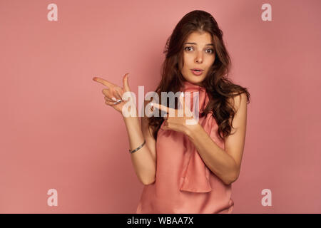 Eine dunkelhaarige Mädchen in ein rosa Kleid auf einem rosa Hintergrund steht, zeigt mit ihrem Zeigefinger an der Seite, Augenbrauen anheben Stockfoto