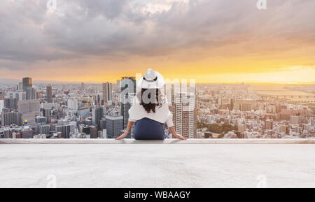Frau in weißen Hut und Kleidung sitzen auf der Dachterrasse, mit Blick auf Osaka City im Sonnenuntergang Stockfoto
