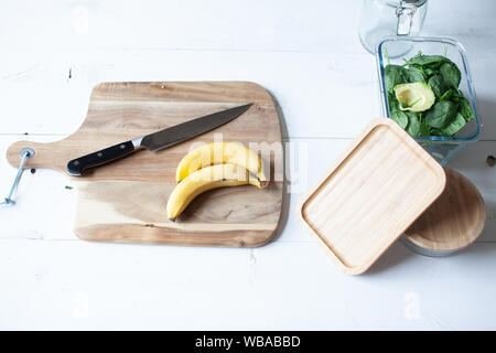 Holz Schneidebrett mit Banane, Spinat und Avocado und ein Messer und Gläser auf weißem Holz- Hintergrund Stockfoto