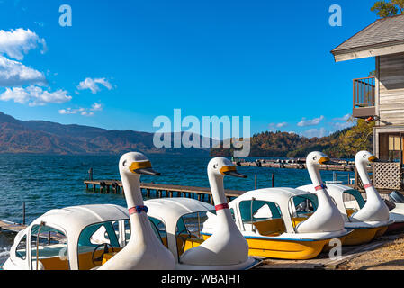 Schönen Herbst Laub Landschaft Landschaften der See Towada in sonniger Tag. Blick vom Lakeside pier, Swan Yachten in den Vordergrund, klarer Himmel, blaue Wasser Stockfoto