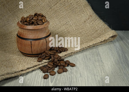 Kaffeebohnen überlaufen von einem kleinen hölzernen Topf neben einem Jute Sack in einem rustikalen noch Leben auf einem Holztisch mit Platz kopieren Stockfoto