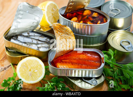 Fisch und Meeresfrüchte, Dosen öffnen Dosen mit sortierten Konserven mit Fisch und Muscheln serviert auf Holztisch mit Petersilie und Pfeffer Stockfoto