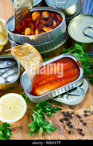 Fisch und Meeresfrüchte, Dosen öffnen Dosen mit sortierten Konserven mit Fisch und Muscheln serviert auf Holztisch mit Petersilie und Pfeffer Stockfoto