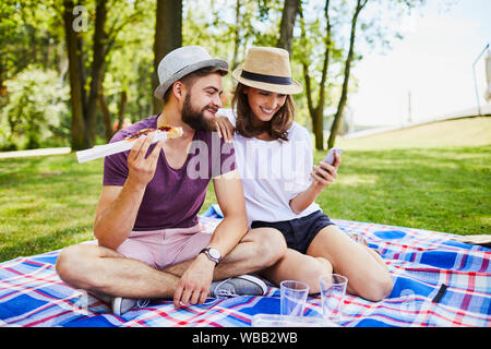 Junges Paar auf Picknick Essen und Suchen am Telefon sitzen im Freien Stockfoto